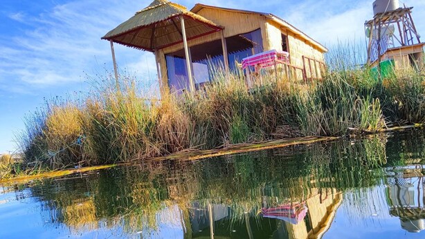 A house with a thatched roof and a water body in front, reflecting the structure. - Uros Quechua Lodge (Puno)