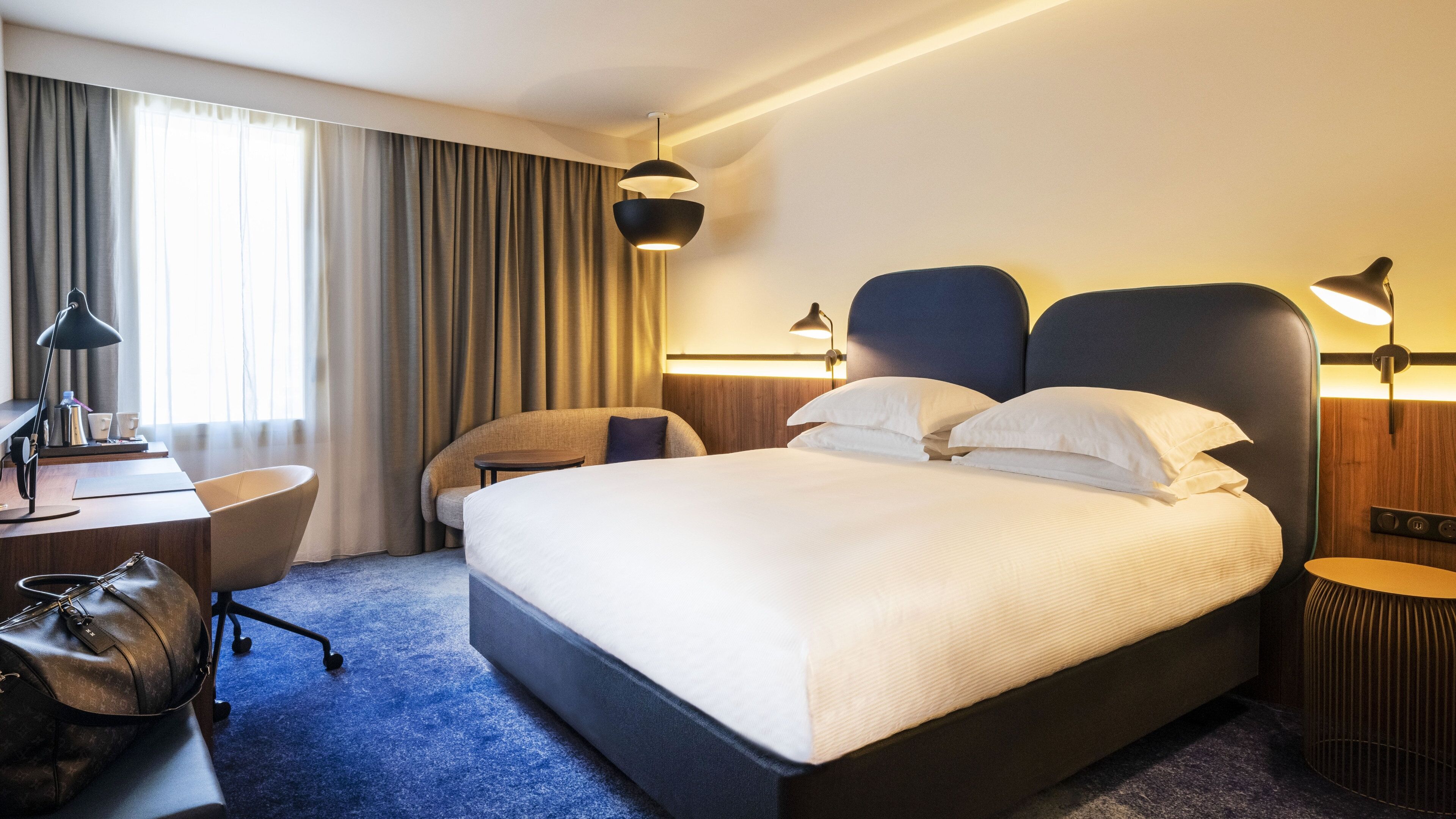 A modern hotel room with a large bed, a desk with a chair, a lamp, and a suitcase.