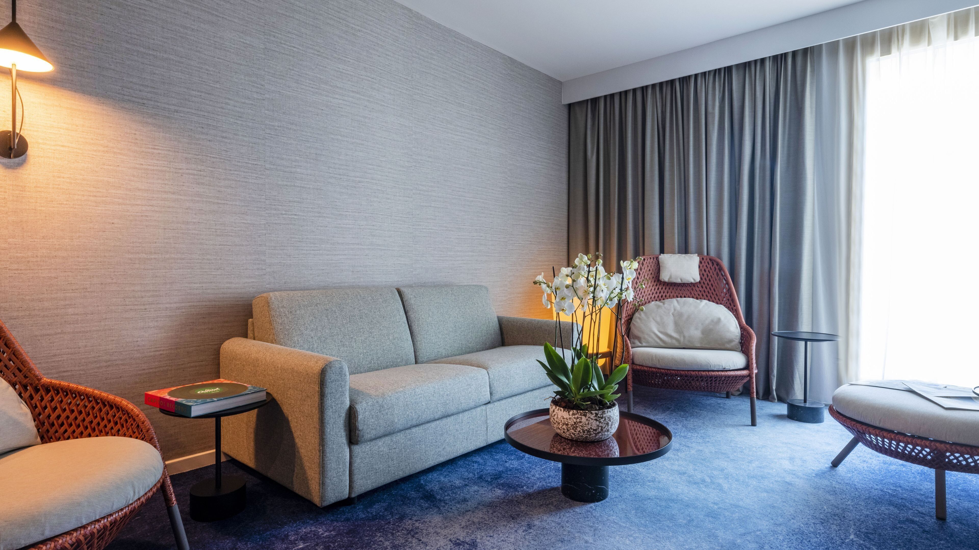 Une chambre d’hôtel moderne avec un canapé gris, un fauteuil en osier, une table basse ronde agrémentée d’une plante et une grande fenêtre avec des rideaux.