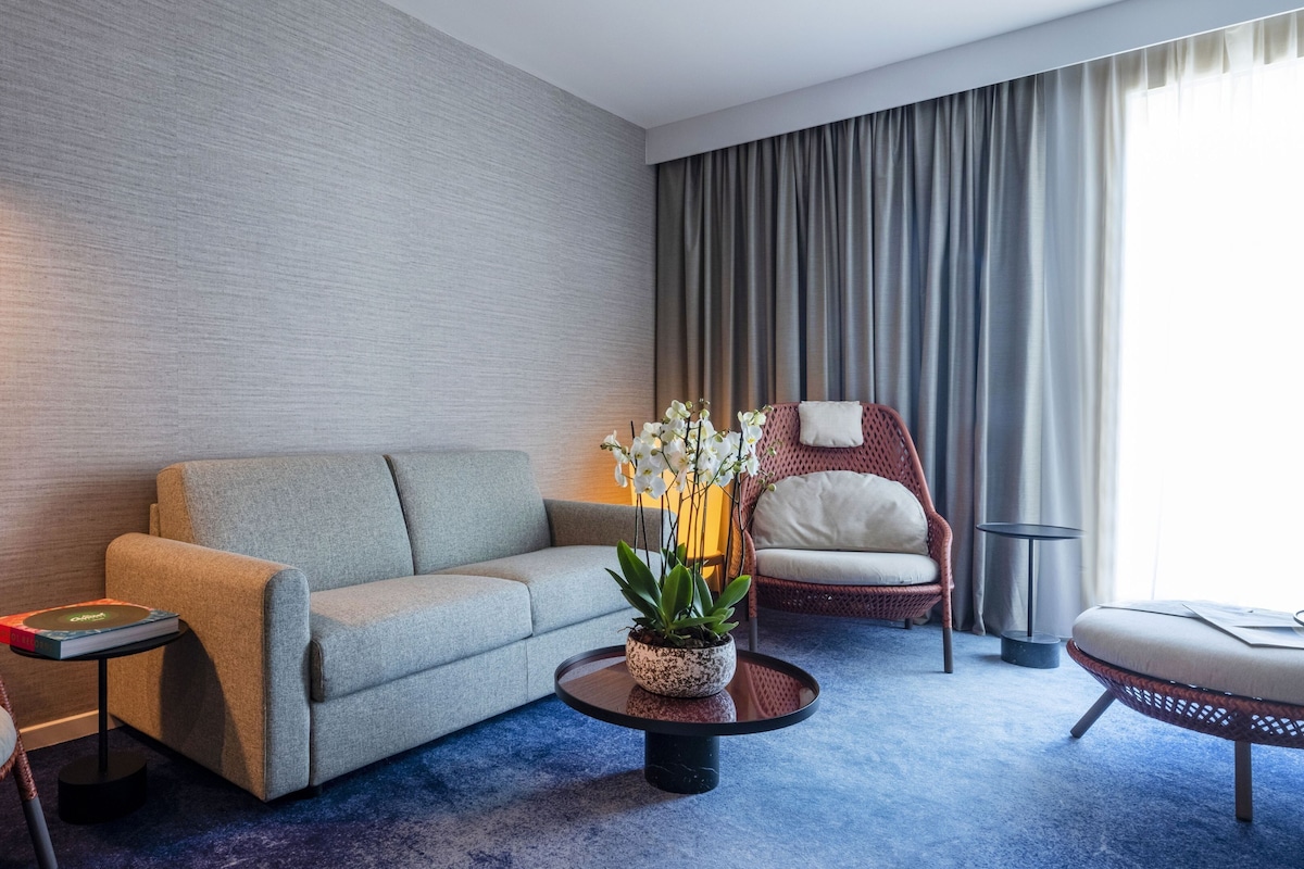 A modern hotel room with a grey sofa, a wicker armchair, a round coffee table with a plant, and a large window with curtains.