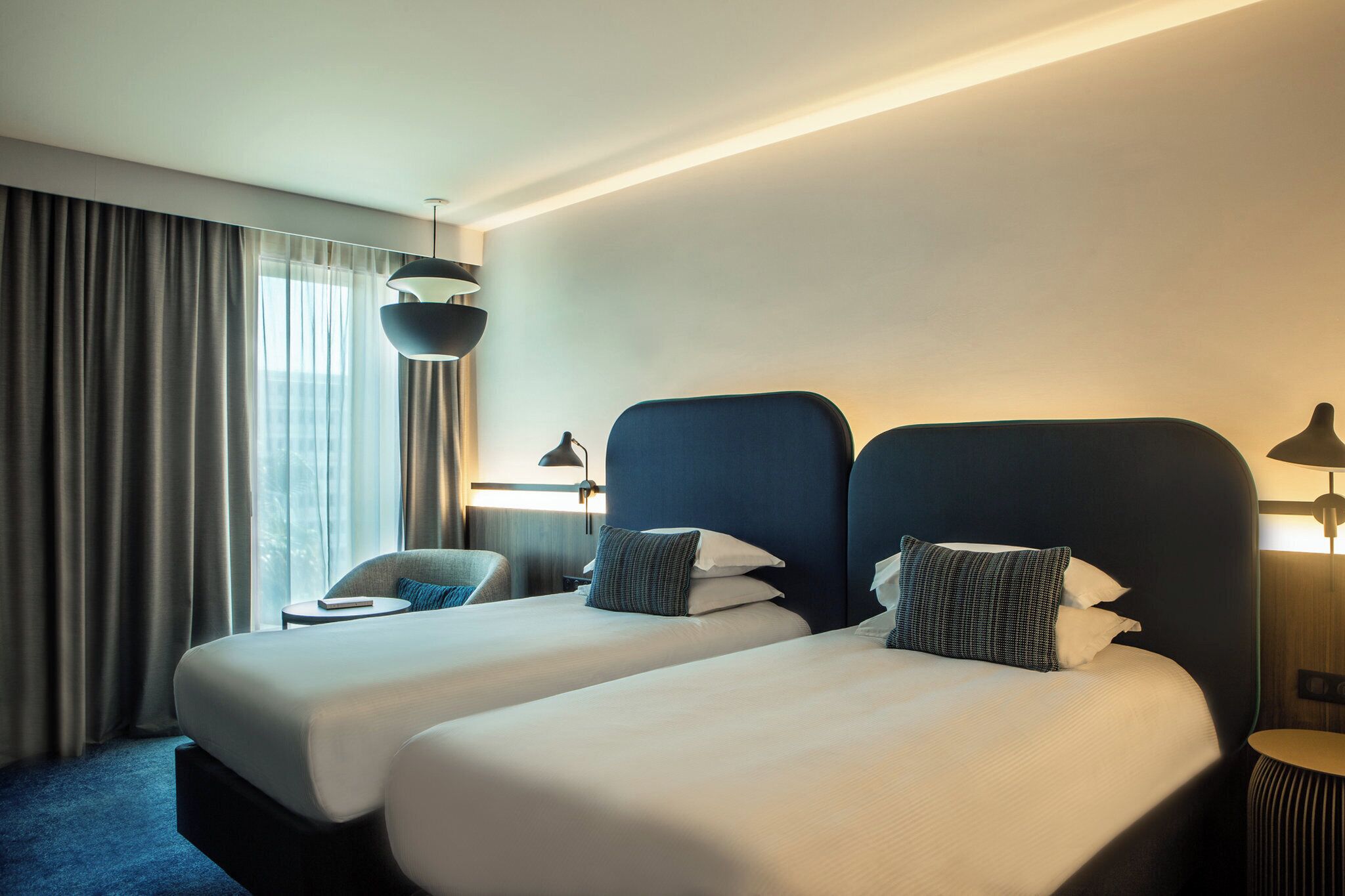 A hotel room with two beds, a chair, a lamp, and a view of the outdoors.
