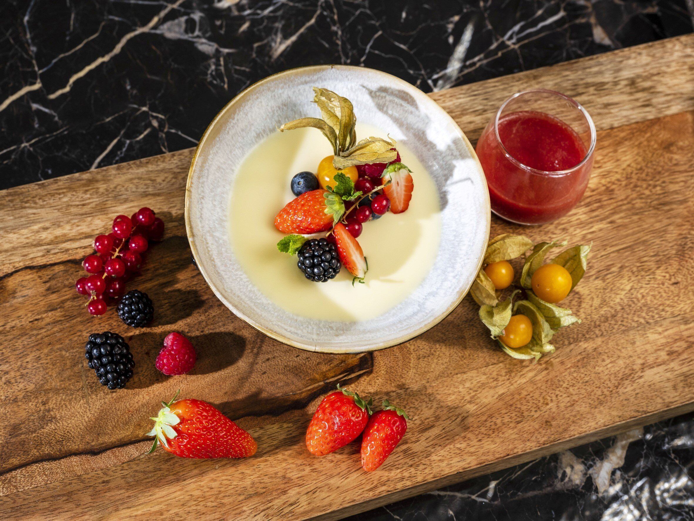 Une assiette de dessert avec divers fruits rouges et un verre d’une boisson rouge, le tout posé sur une planche en bois.