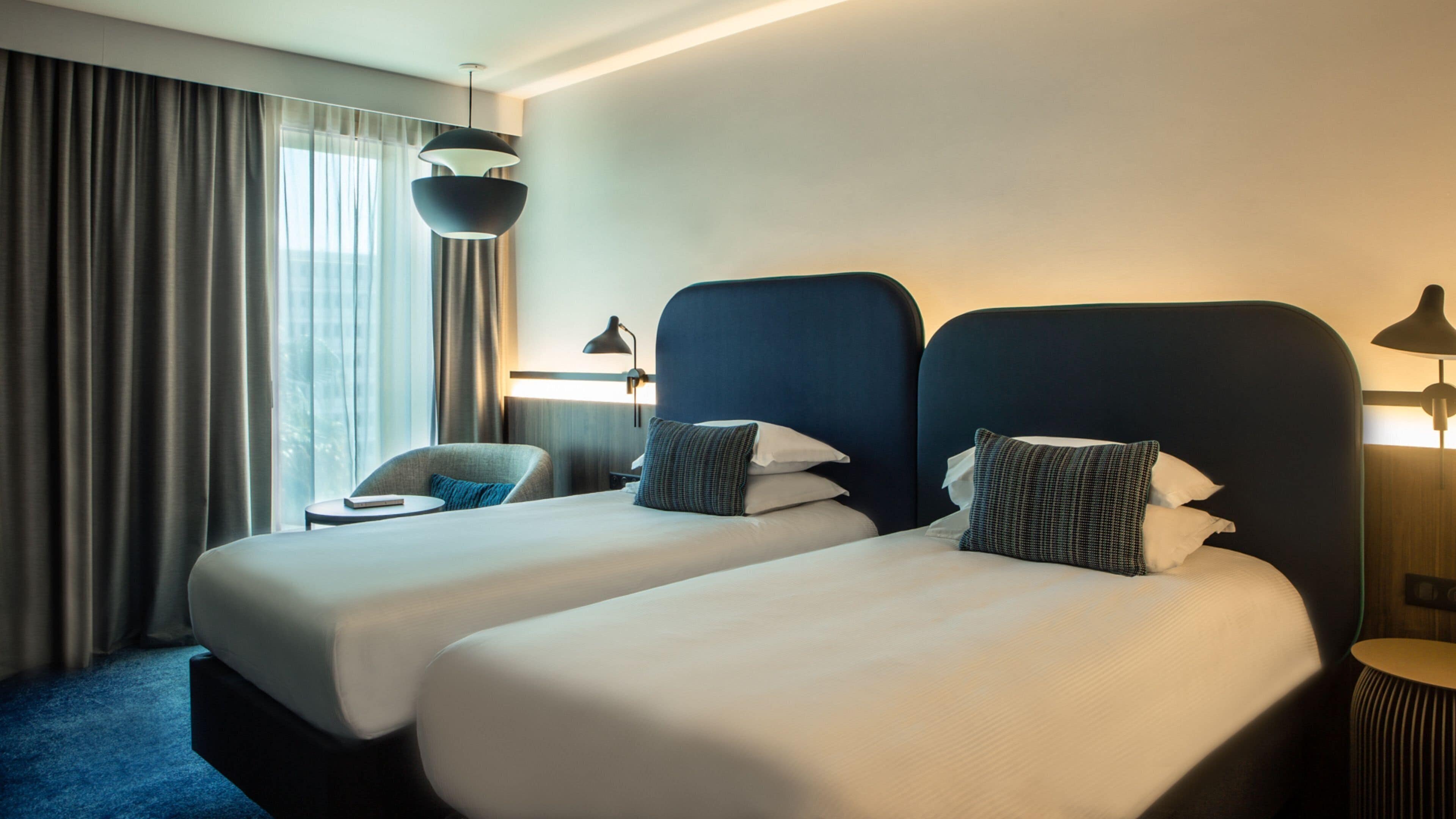A hotel room with two beds, a chair, a desk, and a large window with curtains.