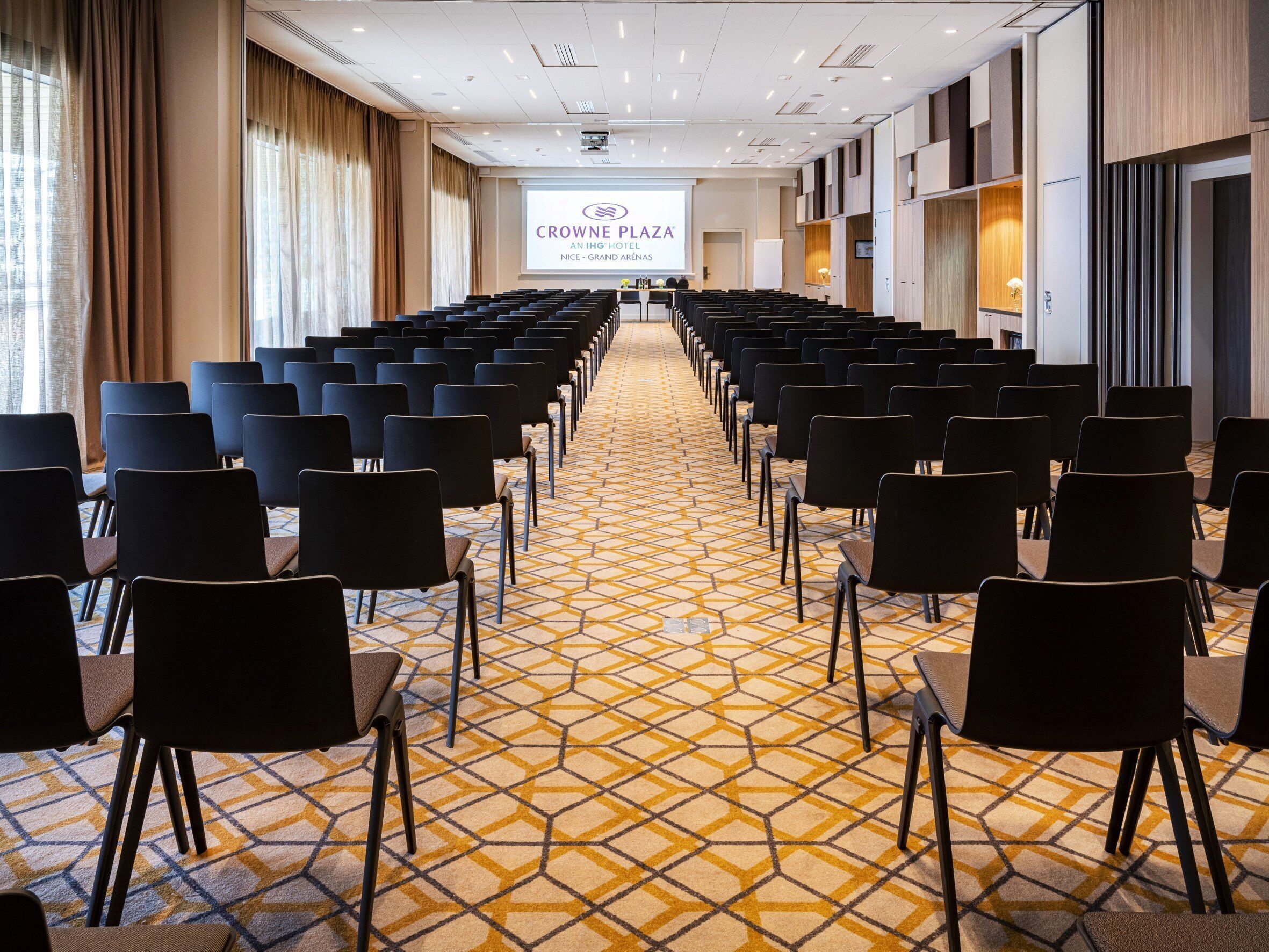 Een vergaderzaal met rijen stoelen gericht op een presentatiescherm, vloerbedekking met een patroon en een modern interieur.