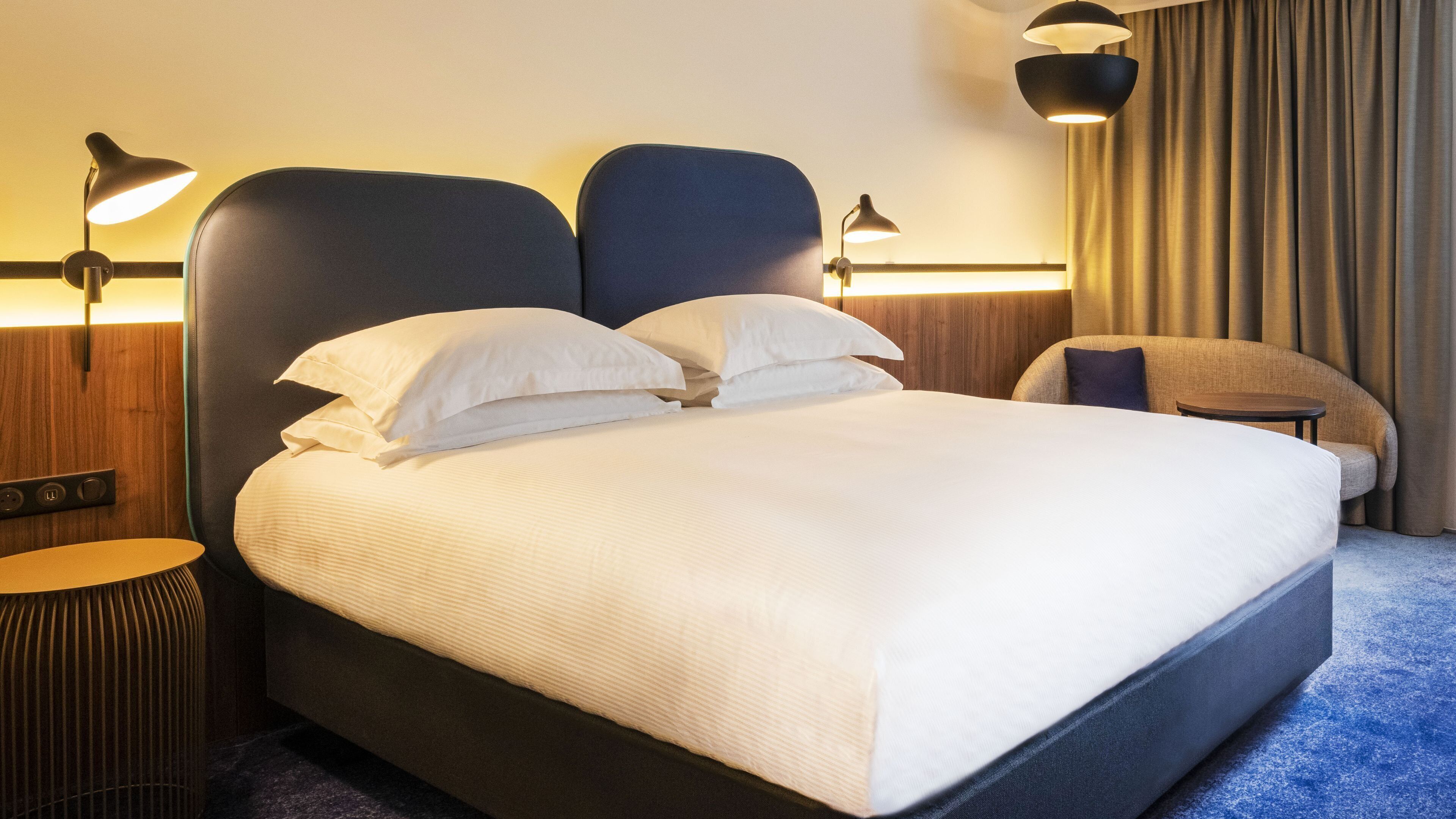 A modern hotel room with a large bed, two bedside lamps, a small table, and a view of the city.