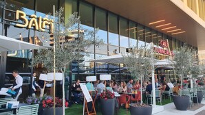 An outdoor seating area with tables and chairs, a menu stand, and a large window with reflections of the sky and surrounding buildings. - Crowne Plaza Nice Grand Arenas by IHG (Nice)