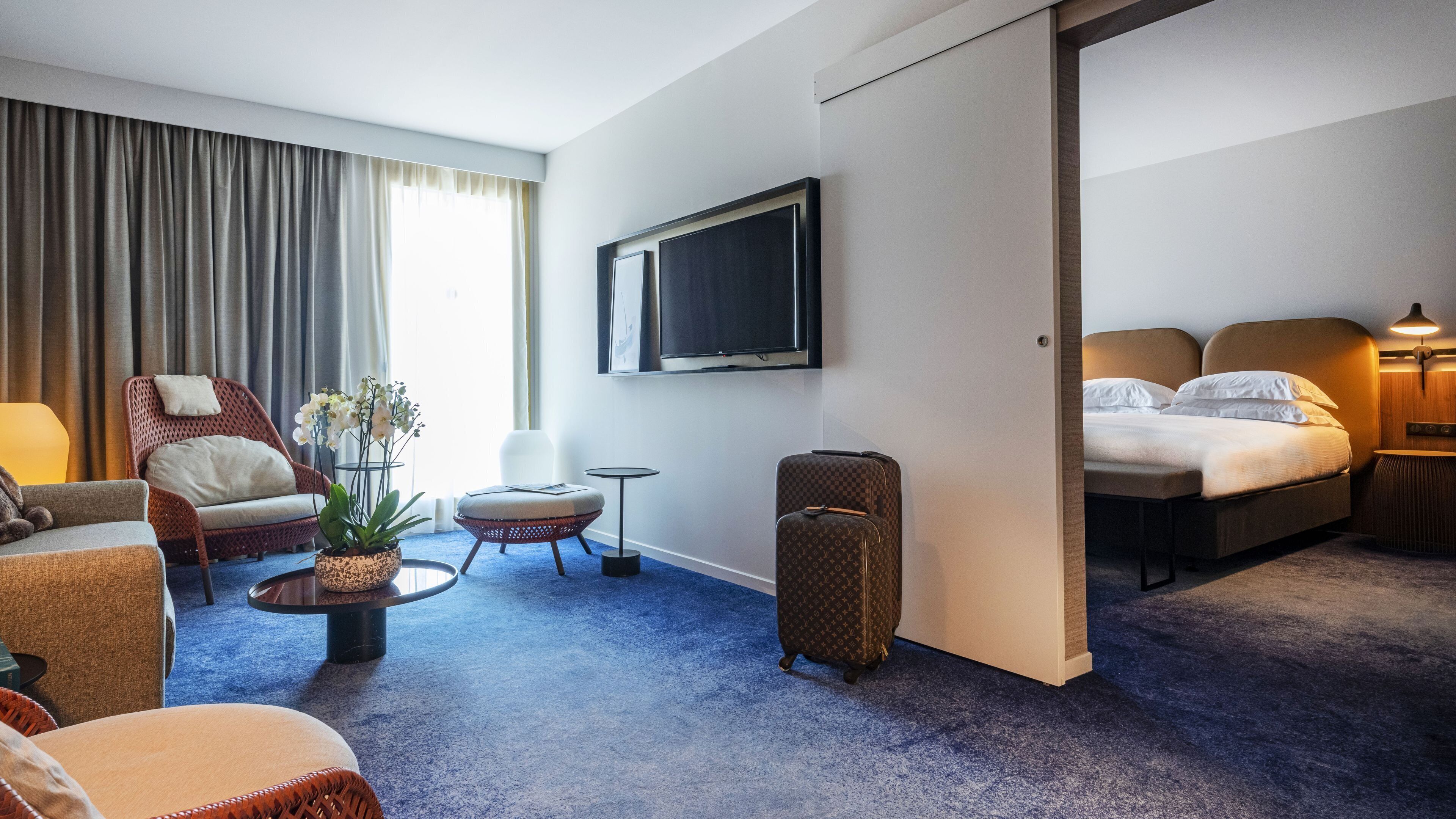 A hotel room with a bed, a TV, a seating area with chairs and a small table, and a large window with curtains.