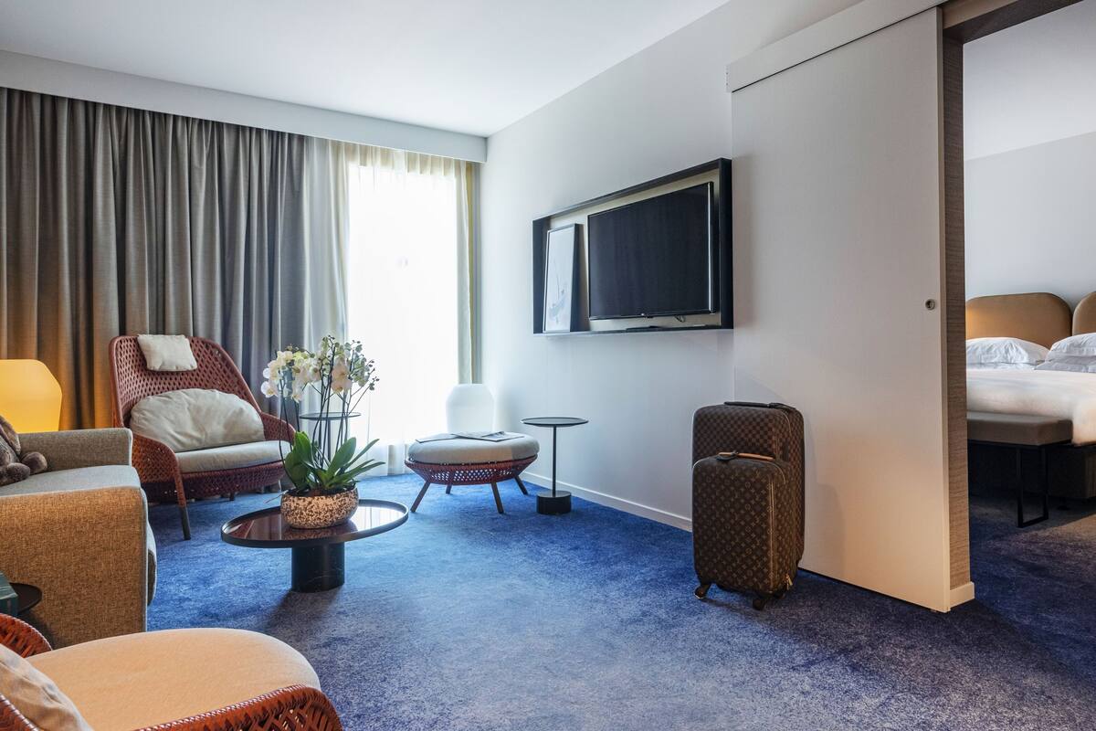 Hotellihuone, jossa on sänky, televisio, oleskelualue tuoleineen ja pienine pöytineen sekä suuri ikkuna, jossa on verhot.
