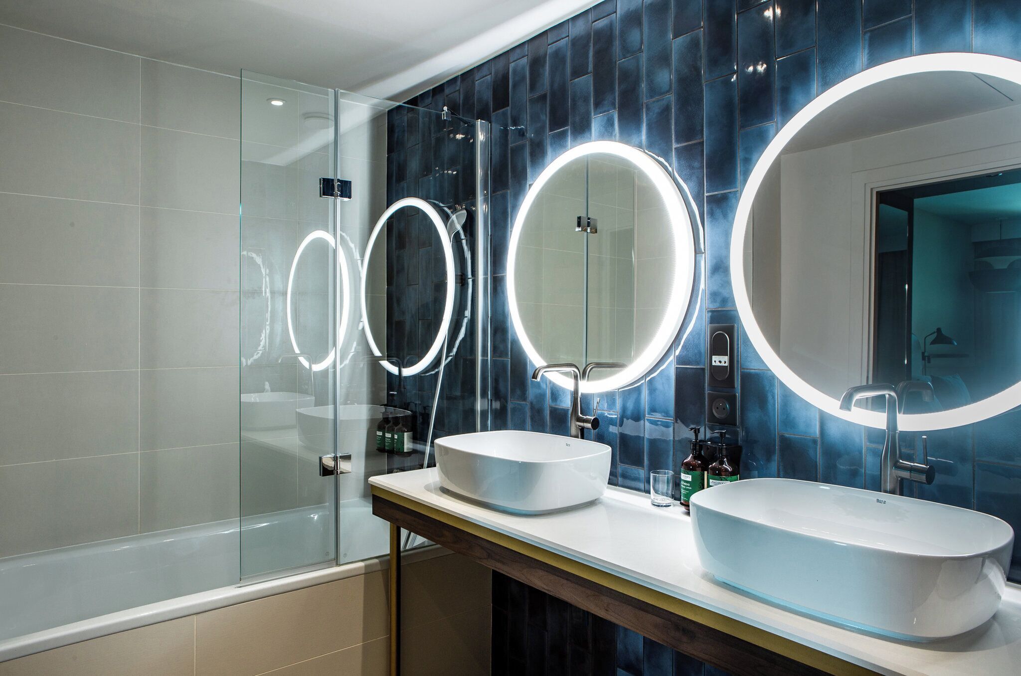 A modern bathroom with two sinks, a glass shower enclosure, and three illuminated mirrors.