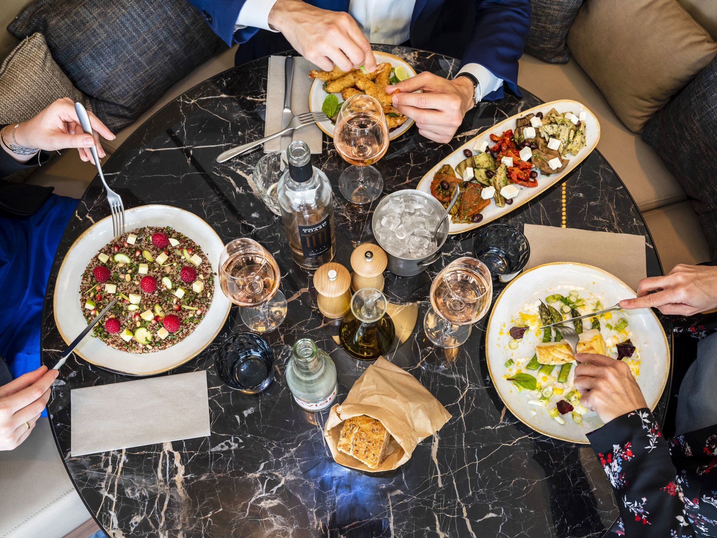 Une table ronde en marbre, sur laquelle on trouve de nombreuses assiettes remplies de nourriture, des verres à vin et des bouteilles d’eau.
