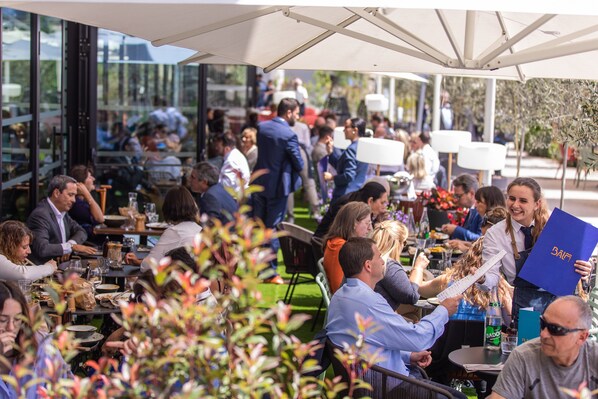 Un área de comedor al aire libre con mesas y sillas, grandes sombrillas y personas cenando.