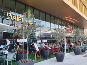 An outdoor seating area with tables and chairs, potted plants, and a building with a large glass facade in the background. - Crowne Plaza Nice Grand Arenas by IHG (Nice)