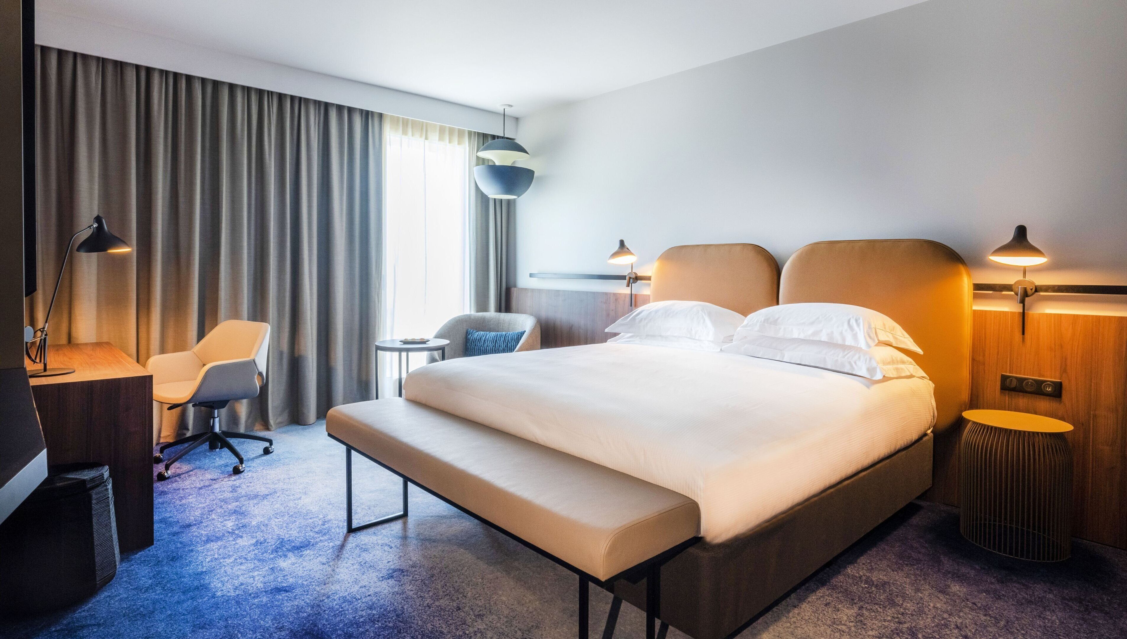 A modern hotel room with a large bed, a desk, a chair, and a small table.