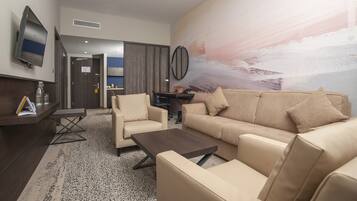 Un salon moderne avec un canapé, des fauteuils et une table basse. Un téléviseur est fixé au mur, il y a un bureau avec une chaise, et un grand tableau abstrait.