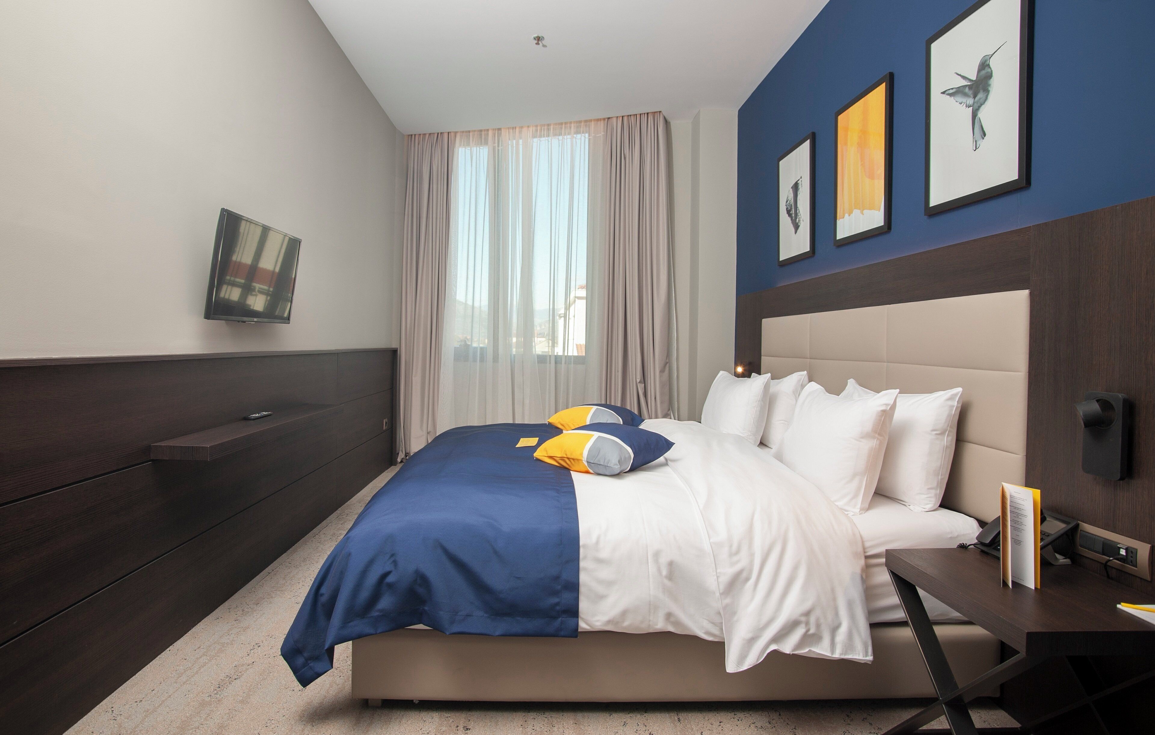 Una habitación de hotel moderna con una cama grande, una mesita de noche con lámpara, un televisor de pantalla plana en la pared y una ventana con cortinas translúcidas.