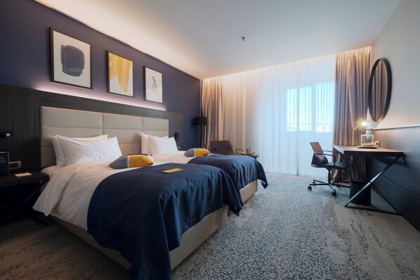 A hotel room with a large bed, a desk, and a chair. There are two paintings on the wall, a window with curtains, and a ceiling light. - voco Podgorica by IHG (Podgorica)