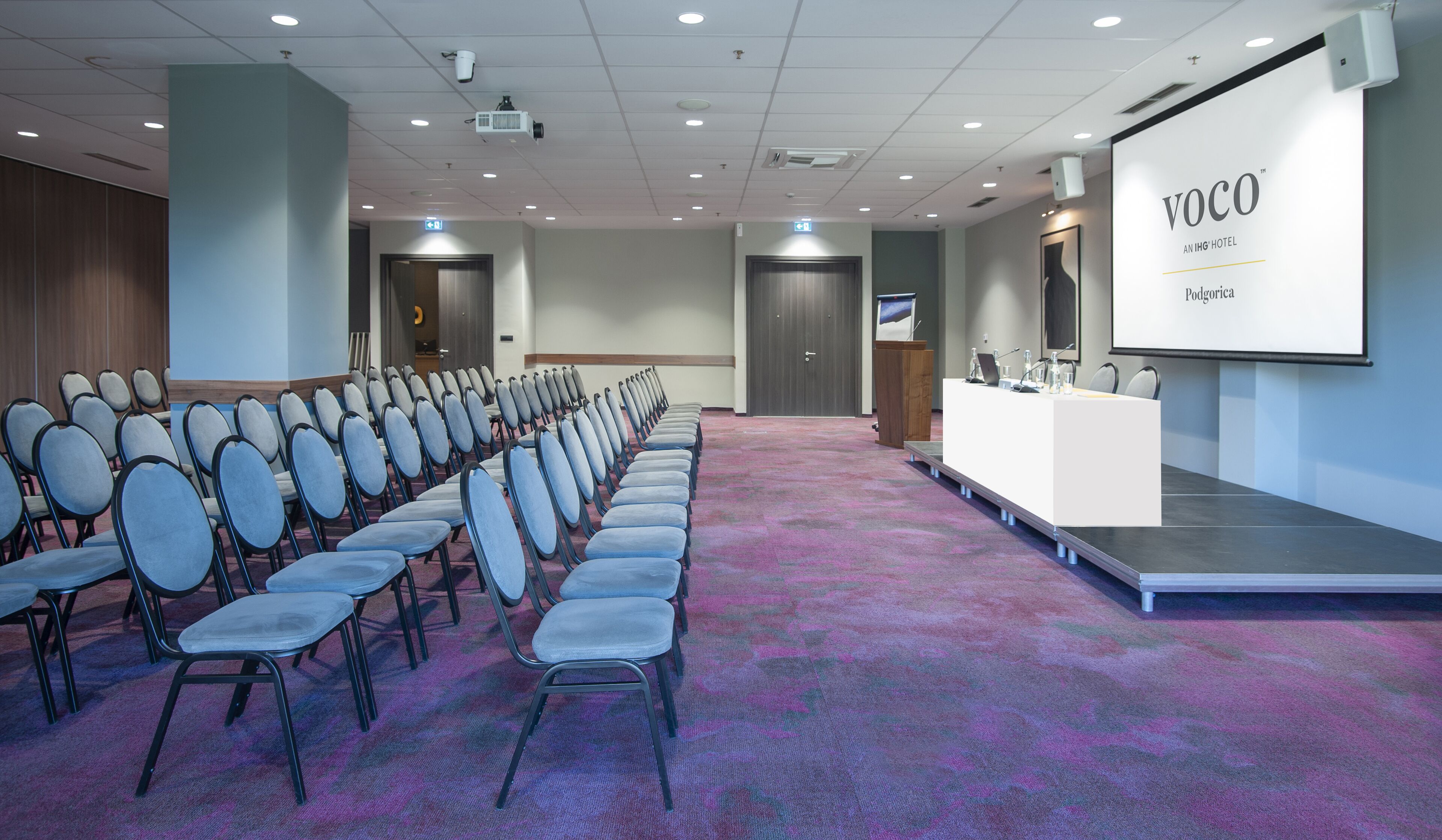 A conference room with rows of chairs, a podium, and a projection screen displaying "VOCO AN IHG HOTEL Podgorica".