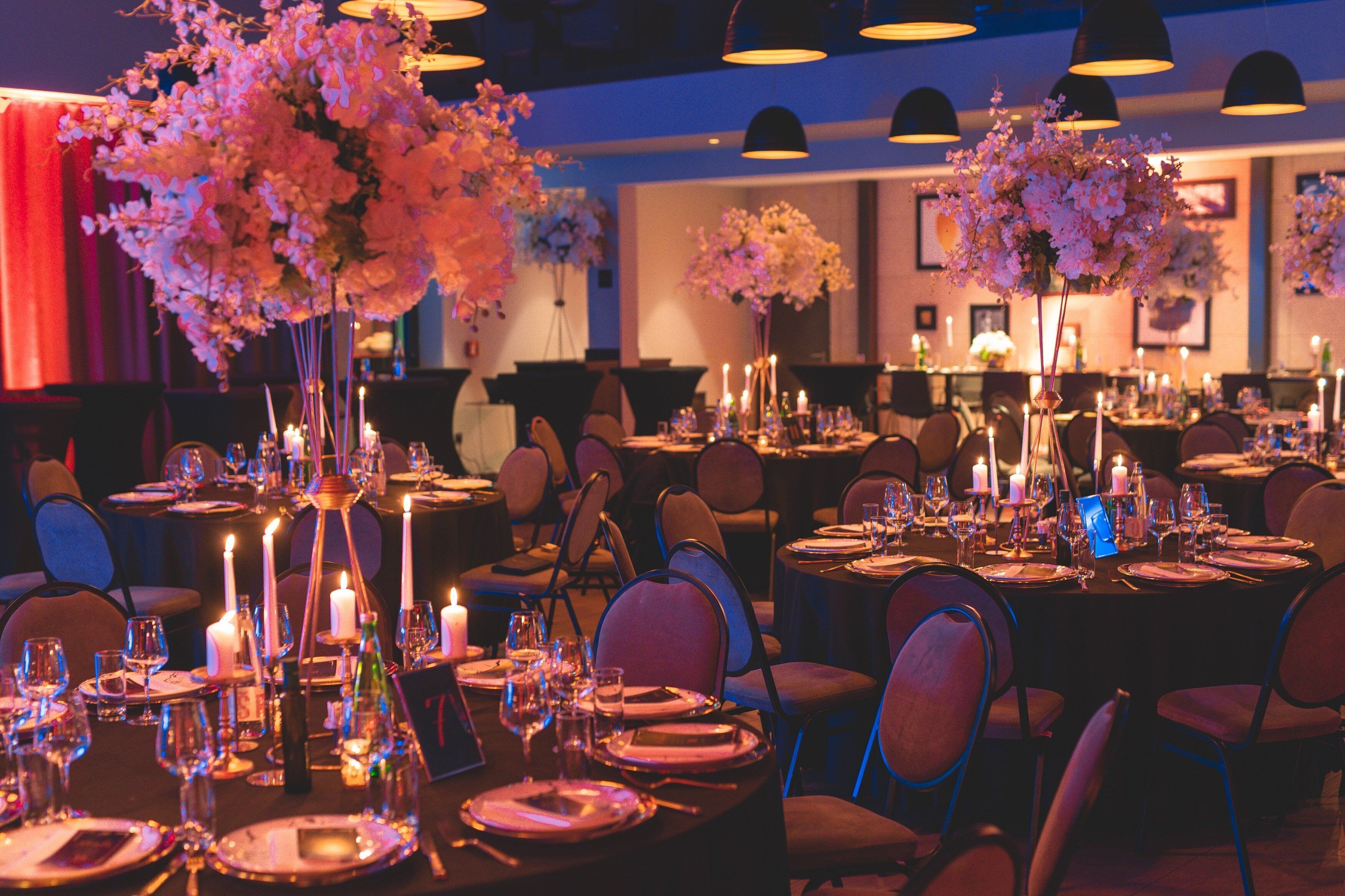 A formal dining room with tables set for multiple guests, featuring floral centerpieces and candles.