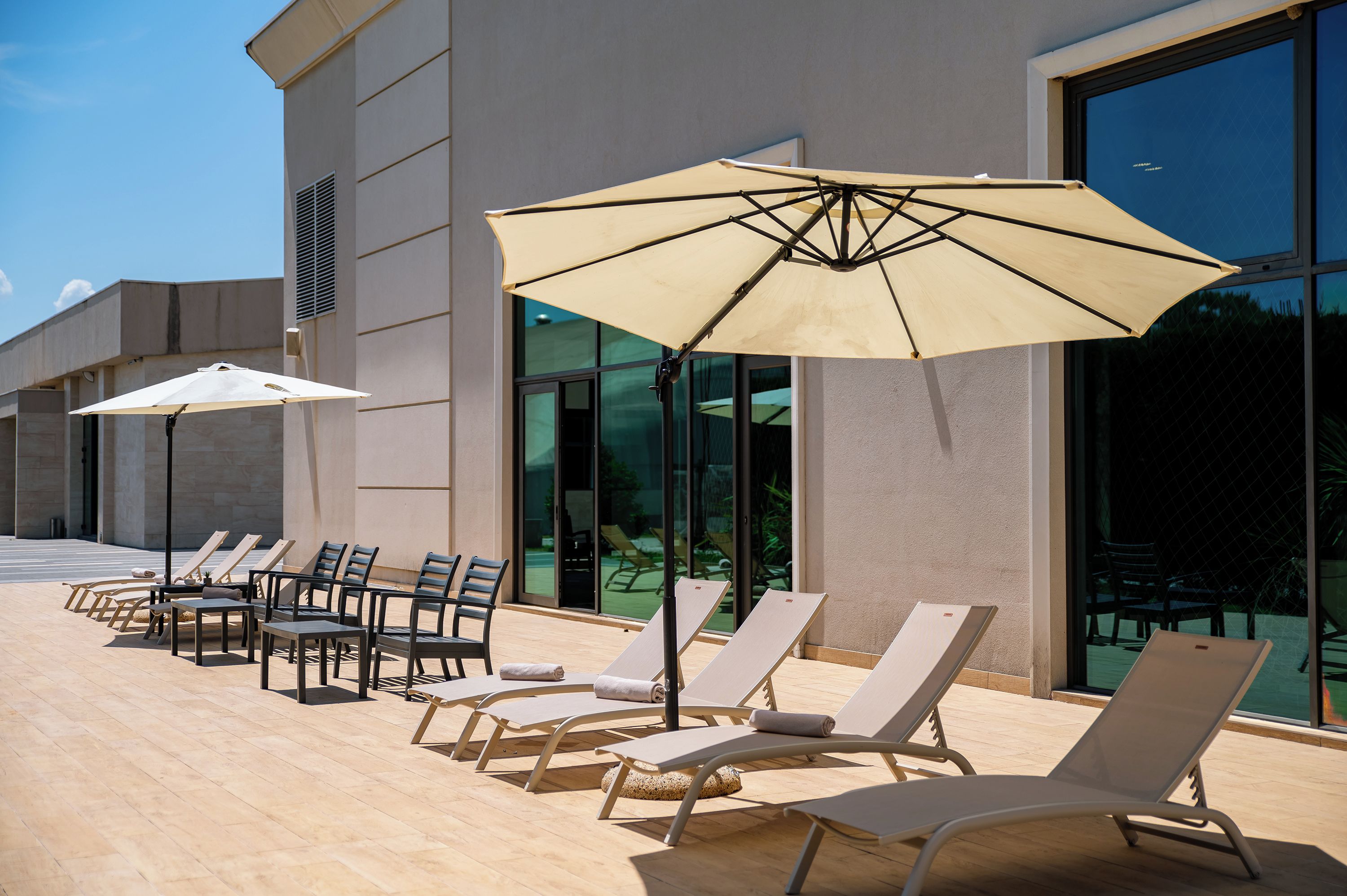 Een zonnig buitenterrein met lounge stoelen en parasols, een zwembad en een moderne gevel.