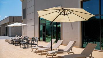 Een zonnig buitenterrein met lounge stoelen en parasols, een zwembad en een moderne gevel.