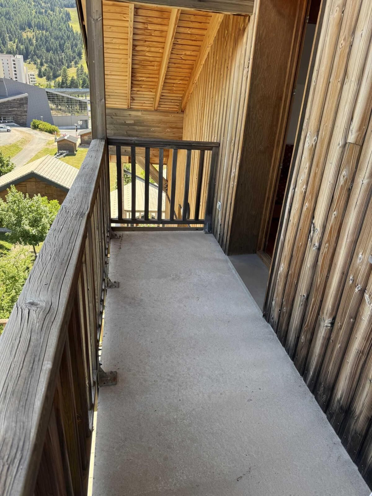 A balcony with a wooden railing, a concrete floor, and a view of buildings and greenery in the background.