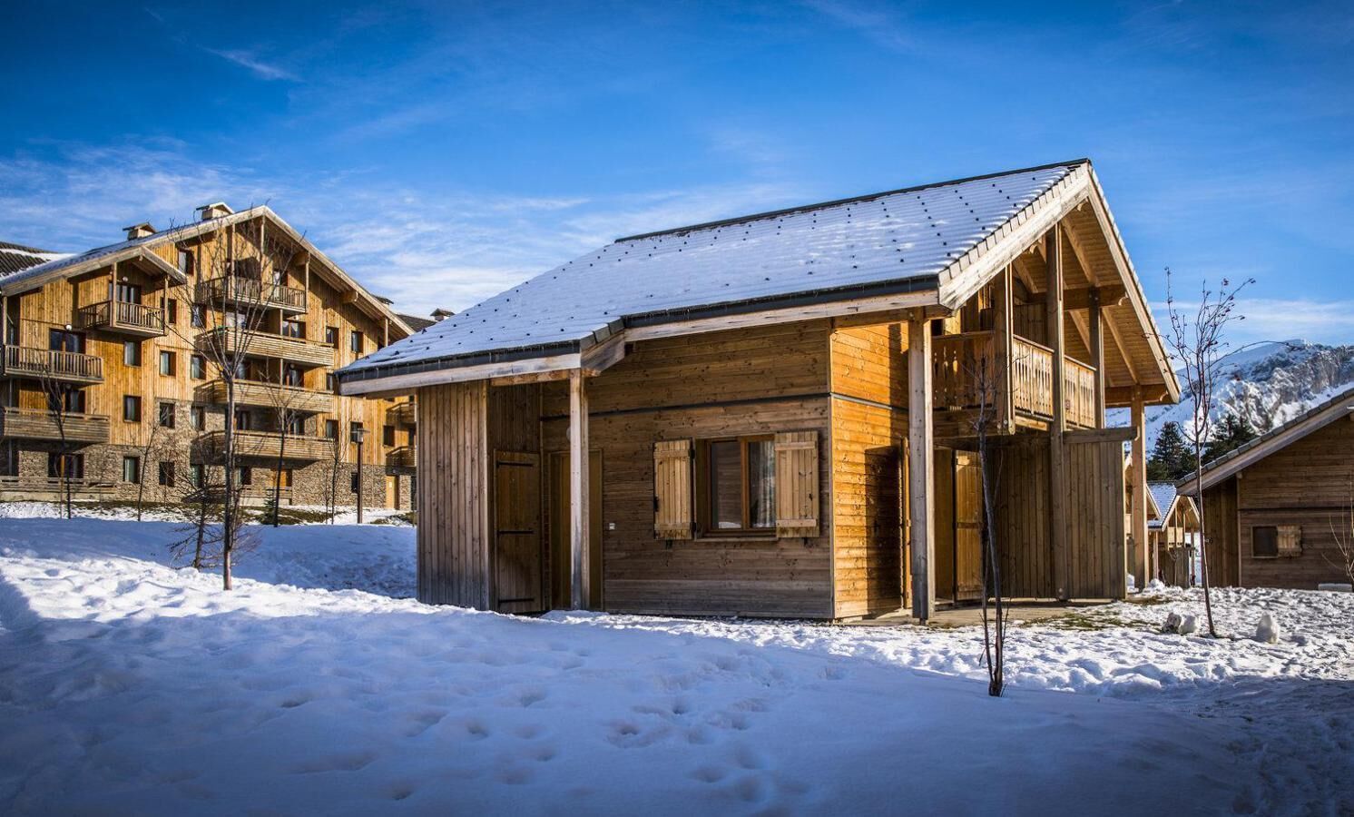 A wooden cabin with a snow-covered roof, surrounded by other wooden buildings, likely part of a resort or lodge.