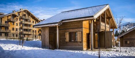 A wooden cabin with a snow-covered roof, surrounded by other wooden buildings, likely part of a resort or lodge.