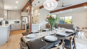 A modern kitchen with stainless steel appliances, a dining area with a black table set for four, and a large chandelier.