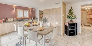 A modern kitchen with a dining area, featuring a wooden cabinet, a small Christmas tree, and a fruit bowl on the table.