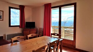 Una habitación con una mesa y sillas de madera, un televisor sobre un mueble bajo, y un balcón con vistas a las montañas.