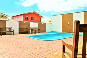 Zona de piscina con suelo de ladrillo, bancos de madera y un edificio rojo al fondo.