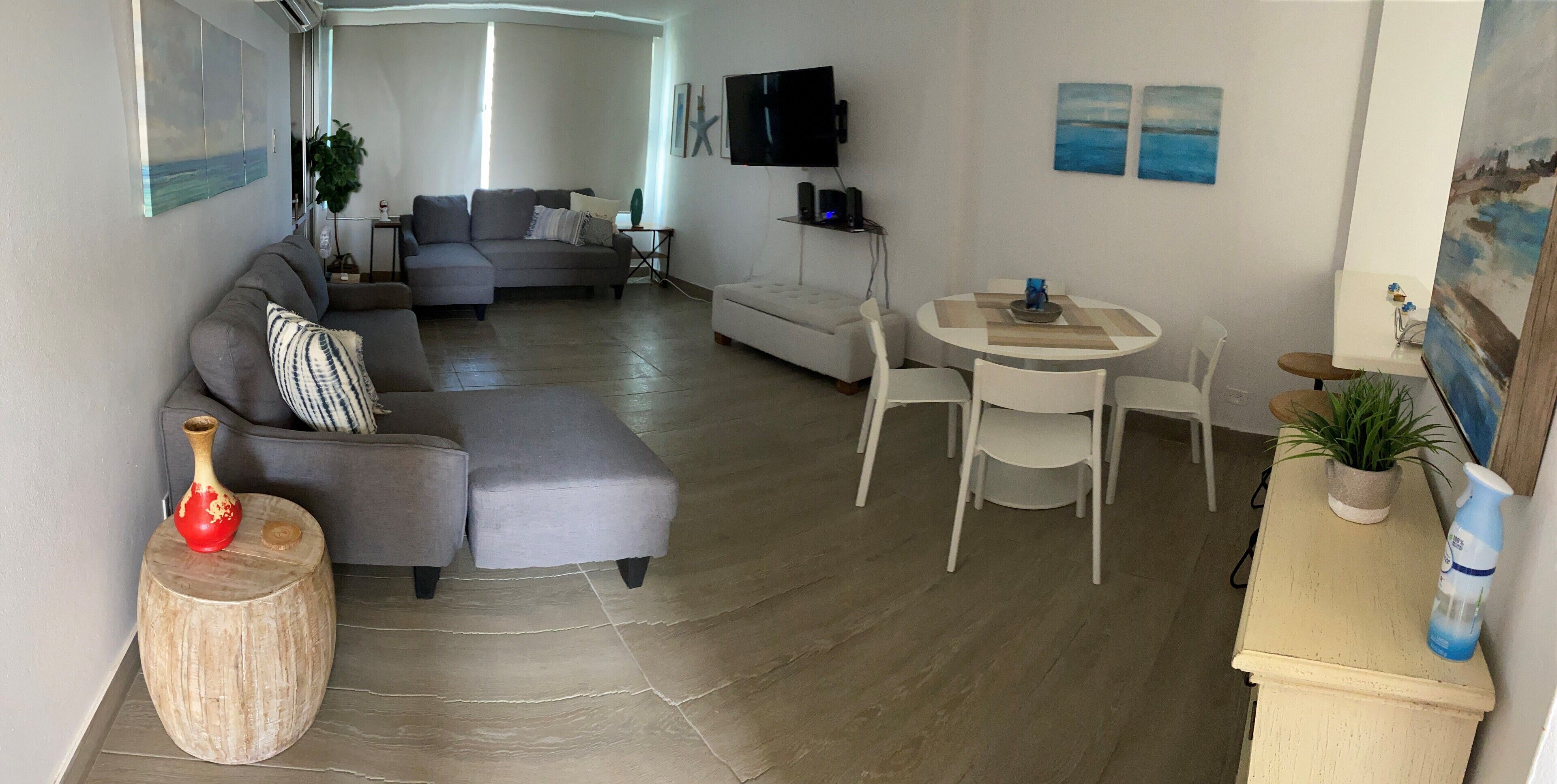Een moderne woonkamer met een grijze hoekbank, een witte fauteuil, een ronde eettafel met vier stoelen en een flatscreen tv aan de muur.