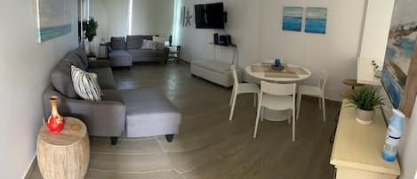 Een moderne woonkamer met een grijze hoekbank, een witte fauteuil, een ronde eettafel met vier stoelen en een flatscreen tv aan de muur.