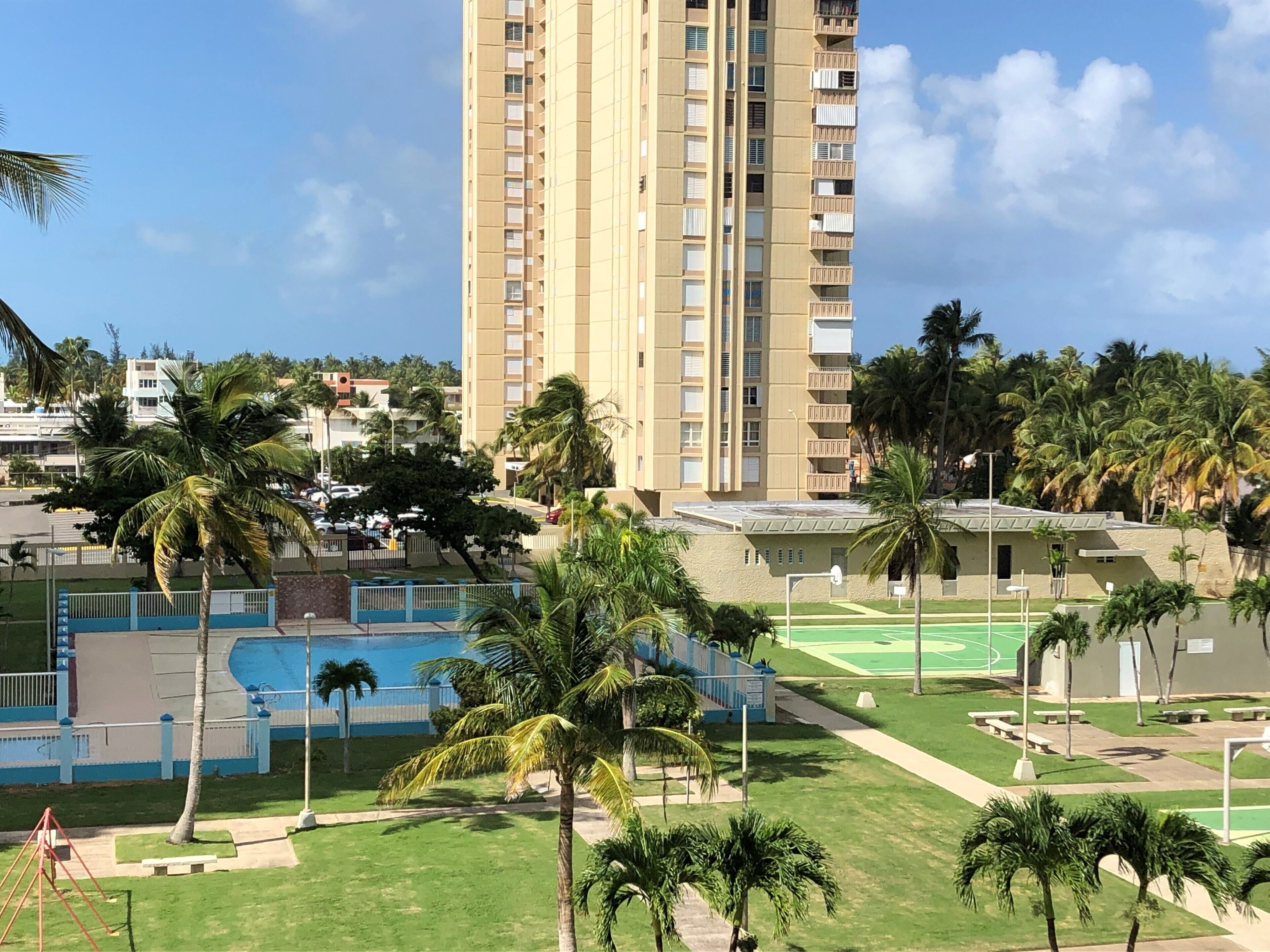 Un edificio alto con varios balcones, rodeado de palmeras y una zona de piscina.
