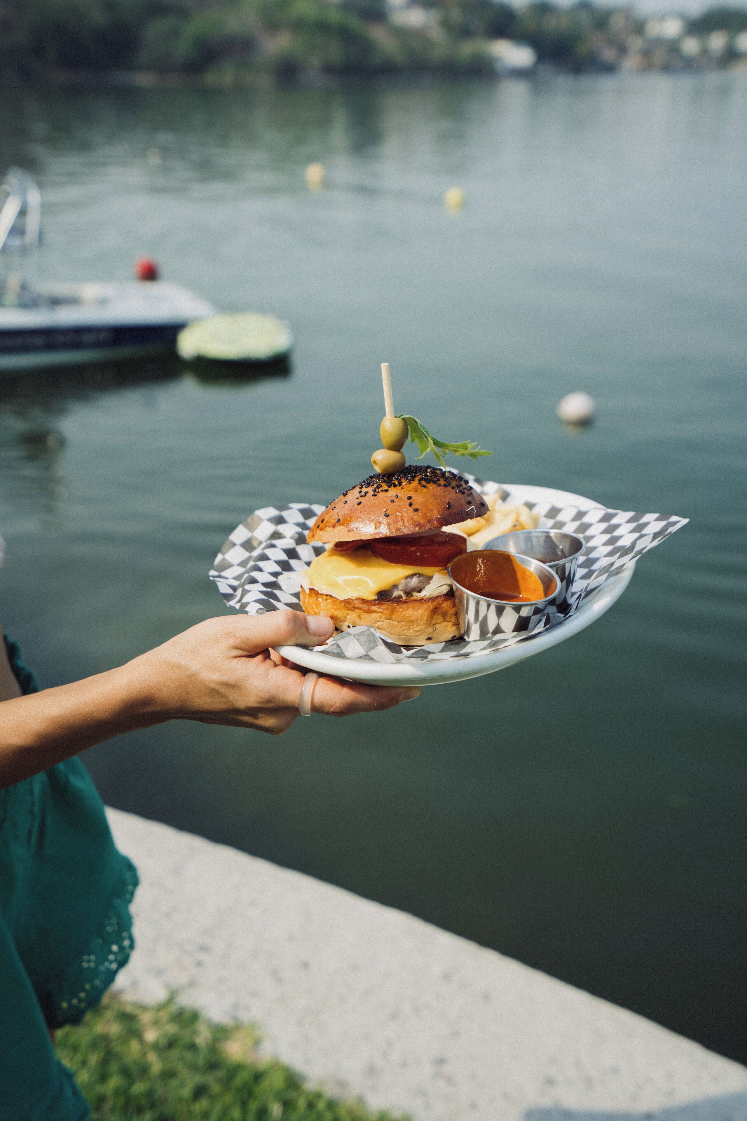 A person holding a plate with a burger and fries by a lakeside.