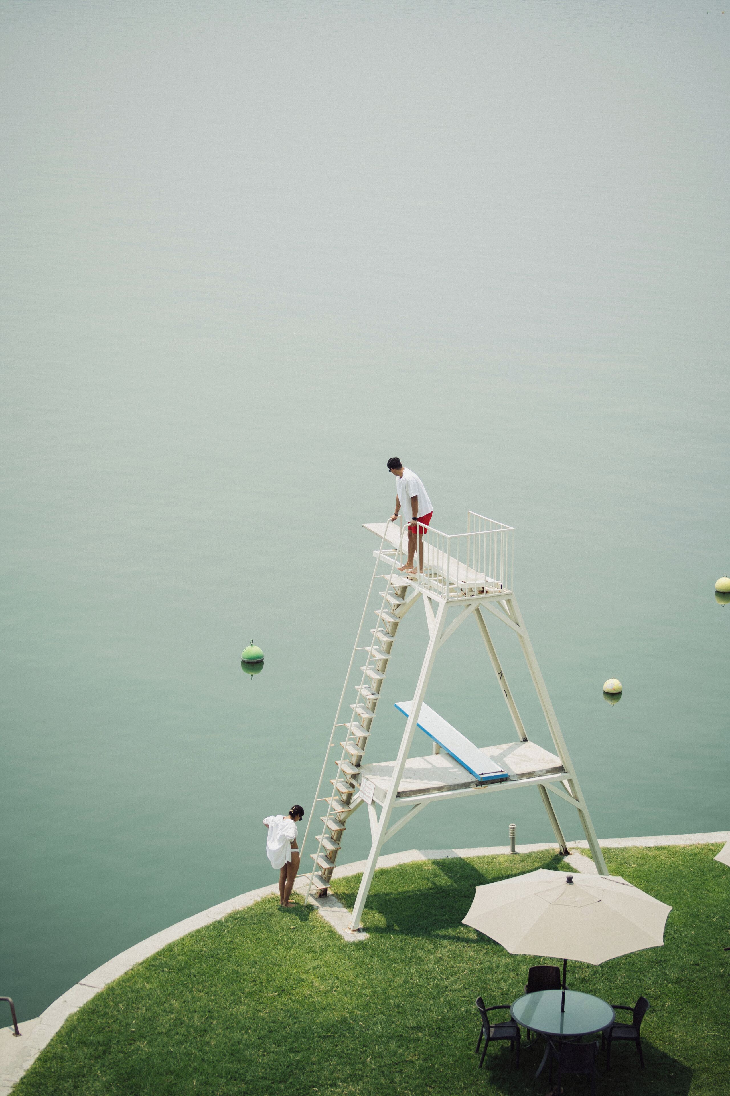 A poolside area with a diving platform, a table with chairs, and a grassy area.