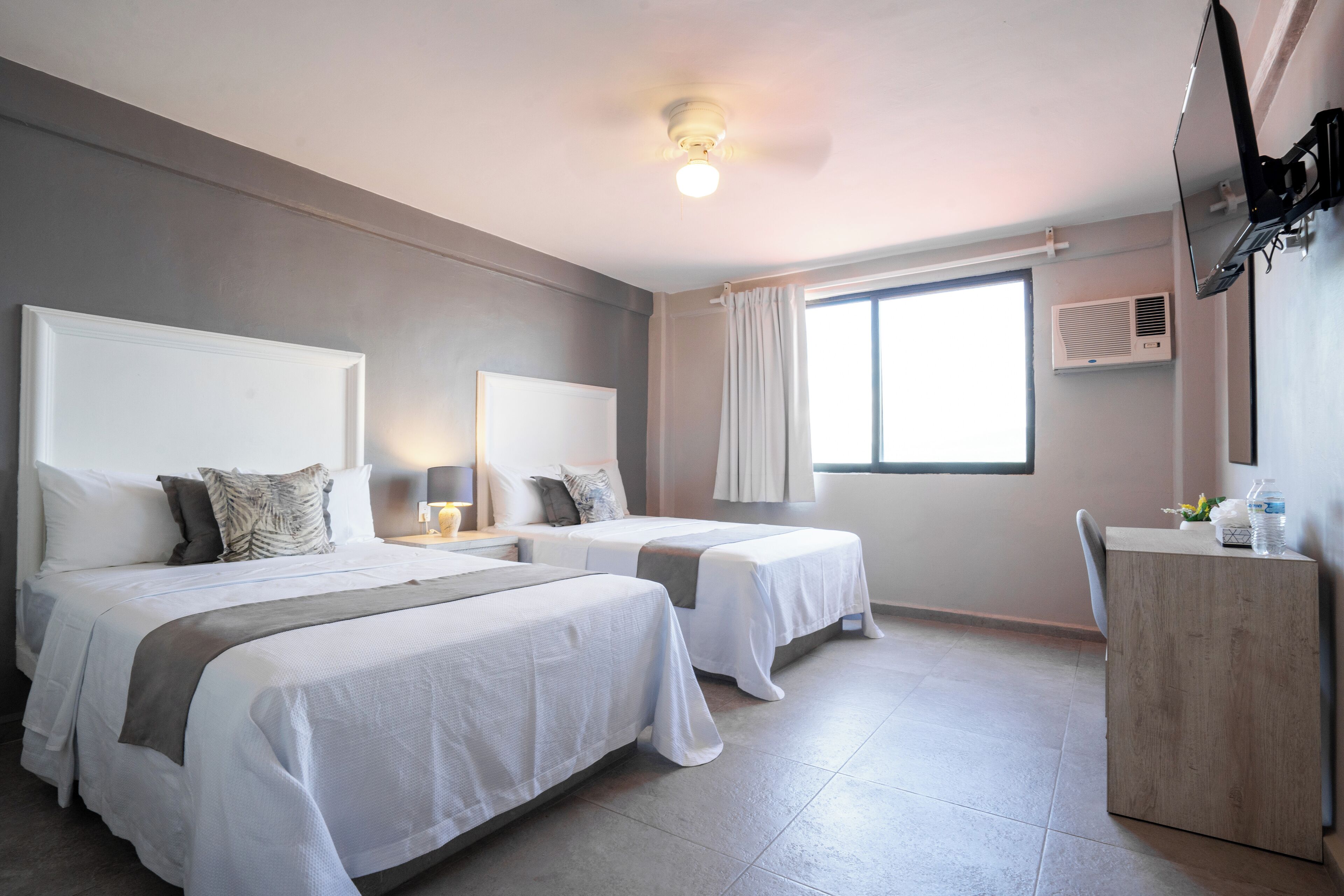 A hotel room with two beds, a desk, a chair, a TV, and a window with curtains.