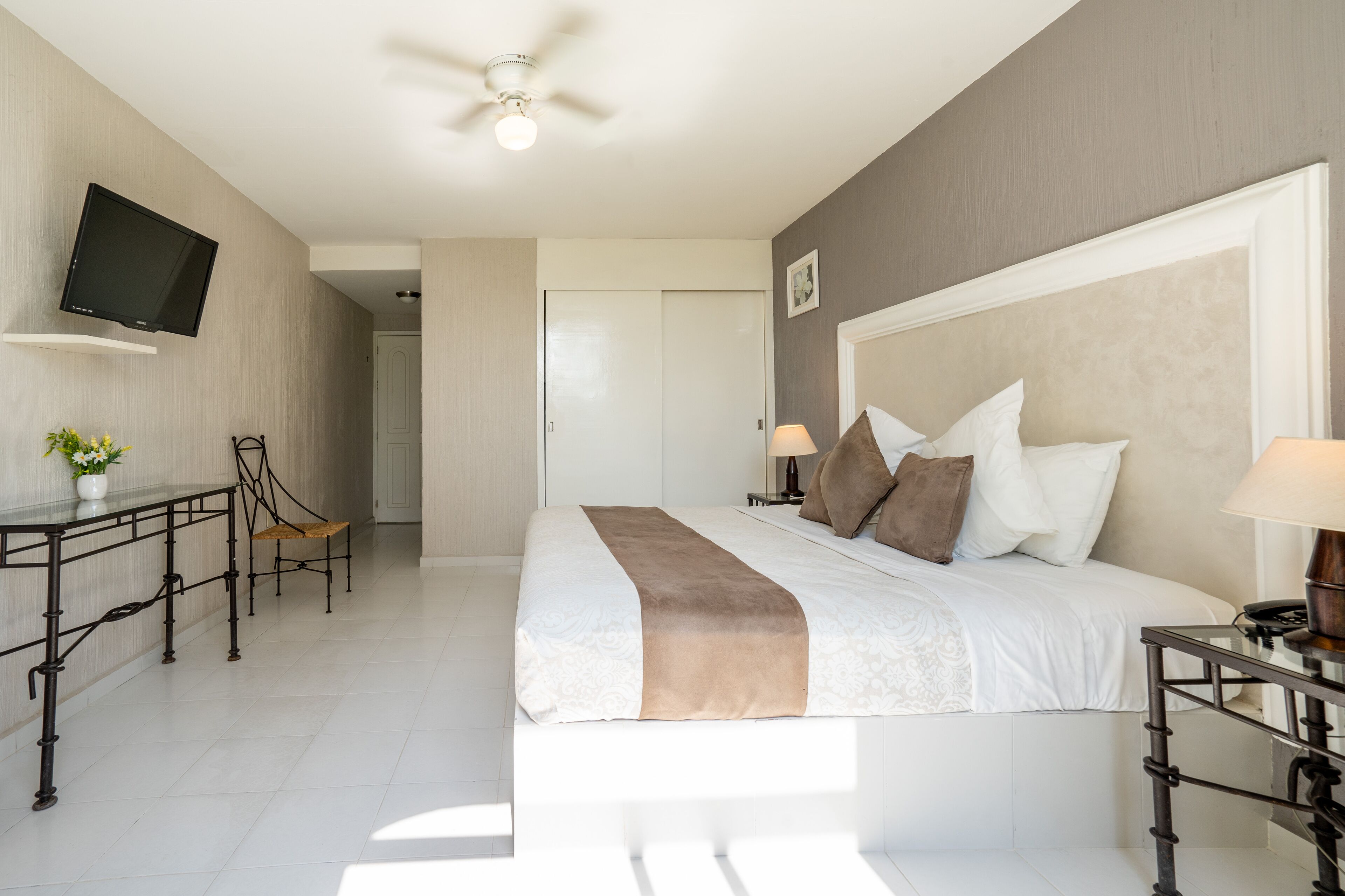 A hotel room with a bed, bedside table, TV, desk, chair, and a ceiling fan.