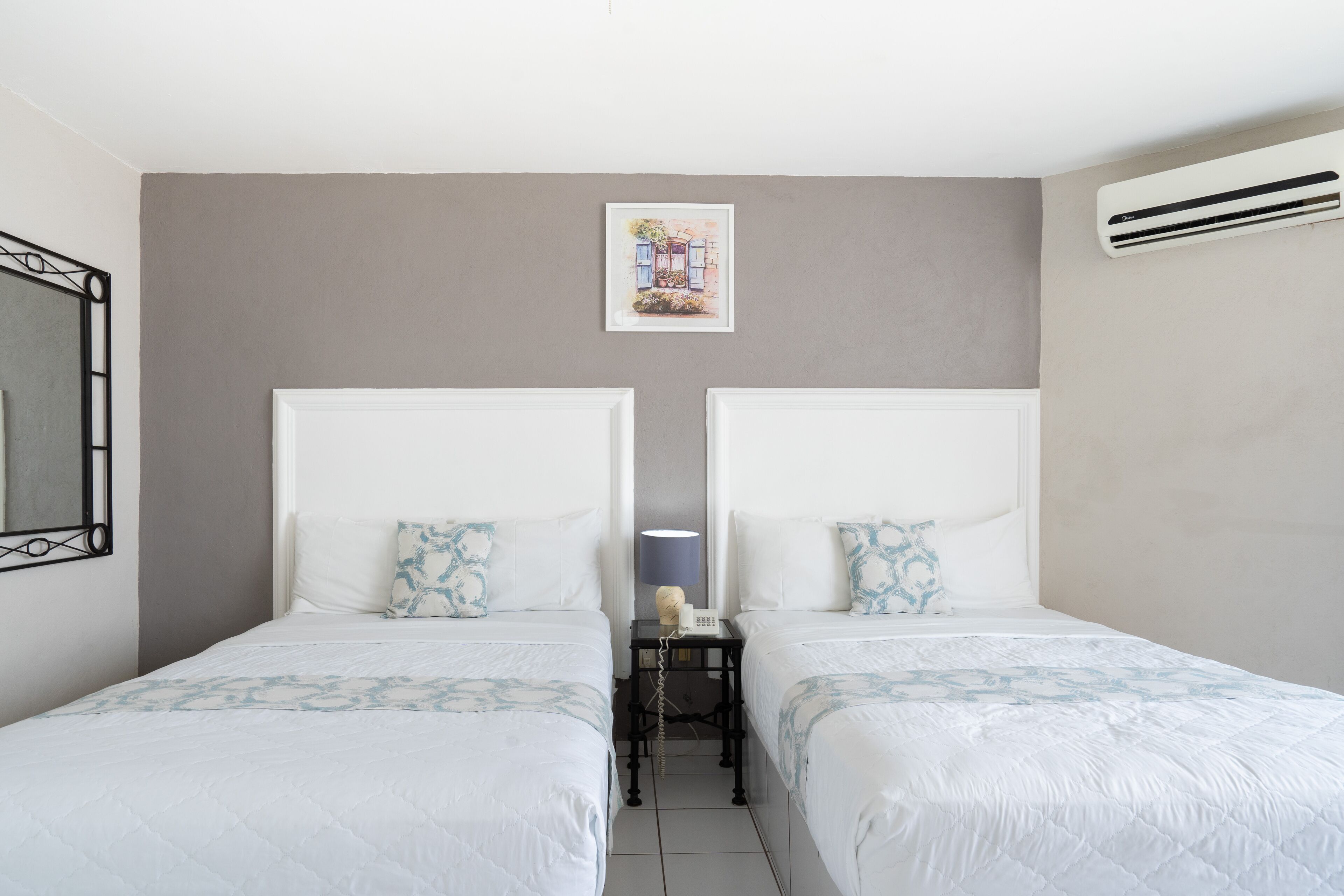 Quarto de hotel com duas camas, uma mesinha com luminária, um quadro pendurado na parede e um aparelho de ar condicionado.