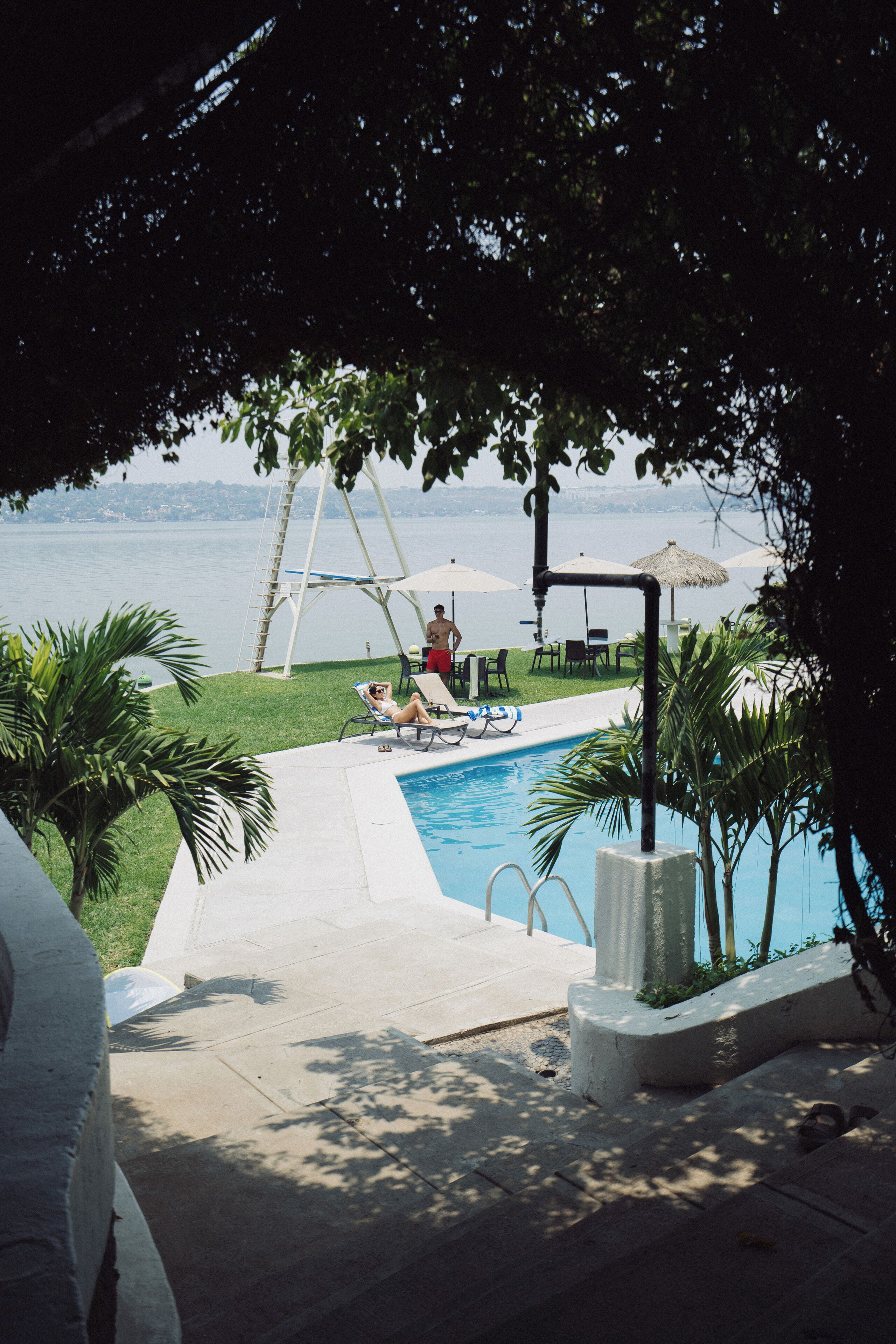 A pool area with a view of the water, lounge chairs, and umbrellas.