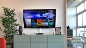 A modern living room with a flat-screen TV displaying a Disney movie, a glass coffee table, a red chair, a potted plant, and a view of the city through large windows. - Rooftop Pool with Ocean and City Views (Santo Domingo)