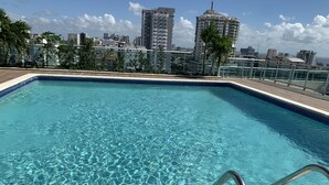 A rectangular swimming pool with clear blue water, surrounded by a tiled deck and metal railings. In the background, there are high-rise buildings and palm trees. - Rooftop Pool with Ocean and City Views (Santo Domingo)