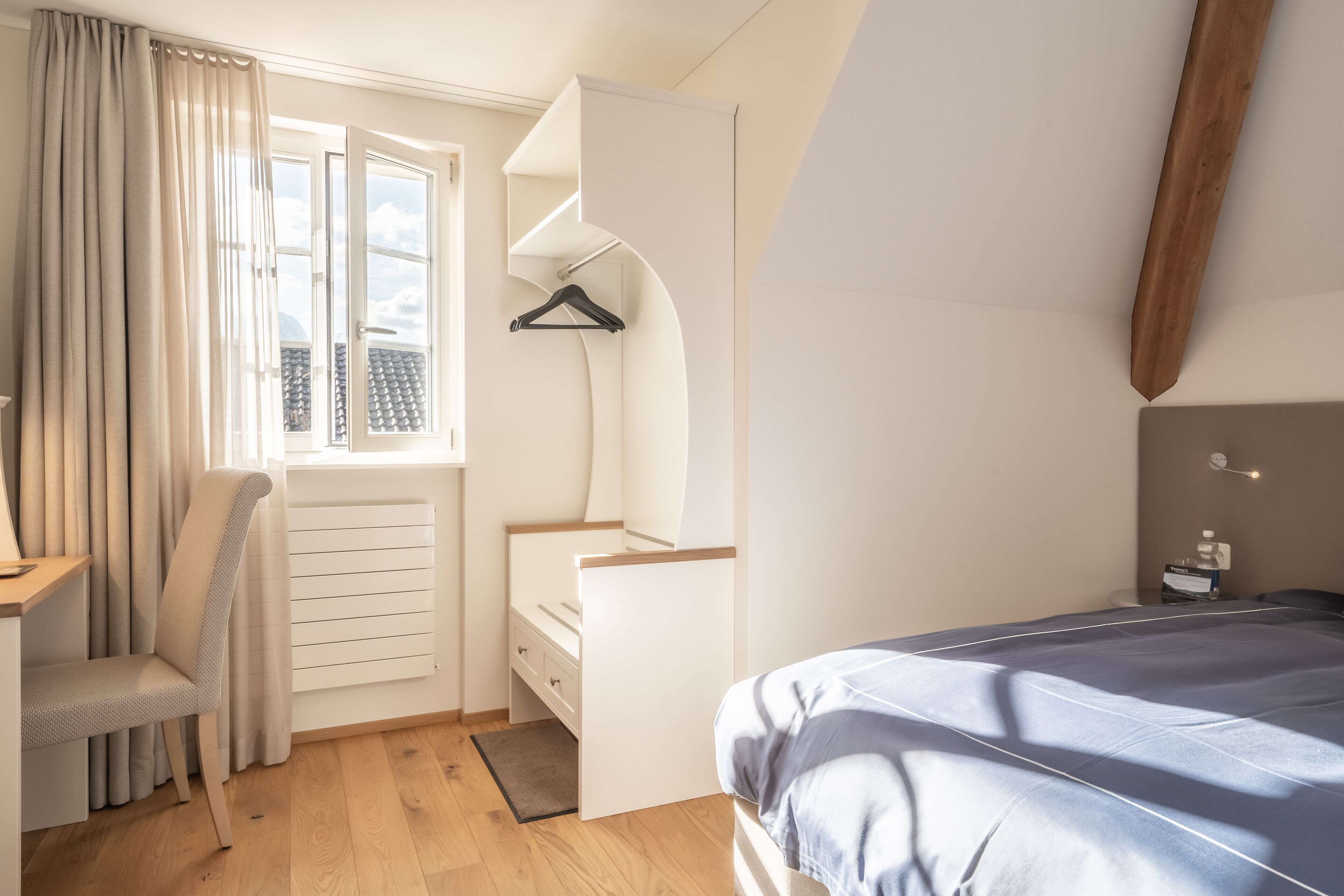 A bedroom with a bed, a desk, a chair, a hanging coat rack, and a window with curtains.
