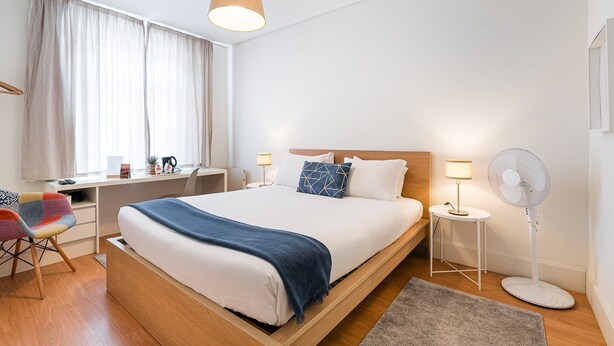 Ein modernes Hotelzimmer mit einem Holzbett, einem Schreibtisch, einem Stuhl, einem Standventilator und einem Fenster mit Vorhängen.