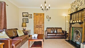 A living room with a brown leather sofa, a wooden coffee table, a wooden door, a fireplace with decorative items, and a chandelier. - 3 bedroom accommodation in Whitby (Whitby)