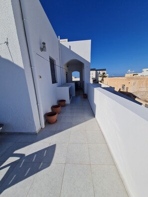 A narrow, sunlit corridor with white walls and terracotta pots, leading to a covered area with a view of the sea.