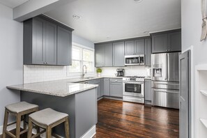 A modern kitchen with grey cabinets, a stainless steel refrigerator, and a central island with stools. - Cozy Downtown Greenwood Retreat - Walkable to Everything (Greenwood)