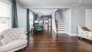 A modern kitchen with stainless steel appliances, a dining area with a tufted armchair and a patterned sofa, and a staircase leading to an upper level. - Cozy Downtown Greenwood Retreat - Walkable to Everything (Greenwood)