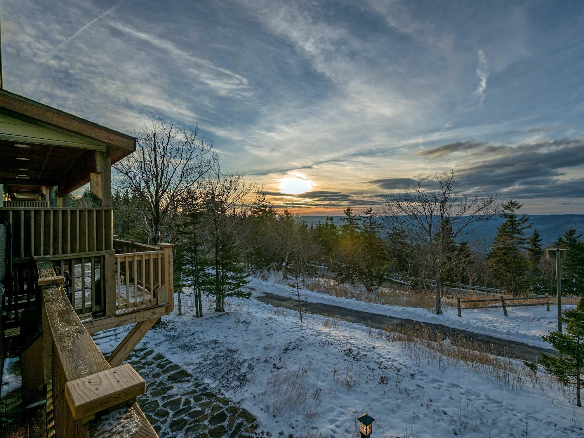 A wooden deck with a stone pathway leading to a cabin, surrounded by snow-covered ground and trees.