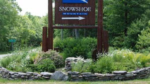 A wooden sign with a blue and white logo for Snowshoe Mountain, an arrow pointing right, and a stone wall with greenery in the foreground. - 5 Br: Black Bear Crossing #4 (Snowshoe)